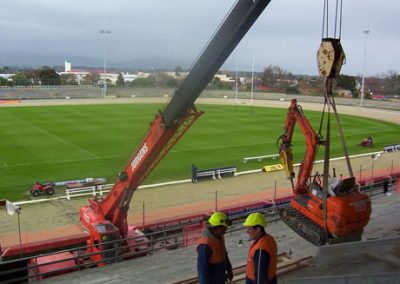 Crane lifting a mini excavator into the Arena Manawatu grandstand above the sports field