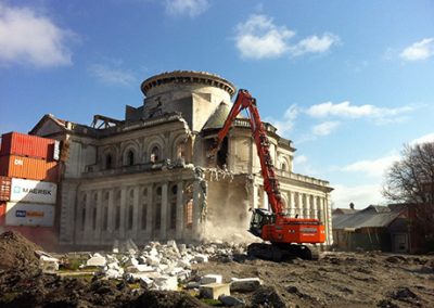 High reach excavator deconstructing the stone facade of the Cathedral of the Blessed Sacrament under a clear blue sky