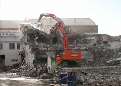 Hitachi excavator demolishing the Makarewa freezing works building with an operator directing from the ground