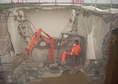 Operator in orange overalls using a mini excavator inside a concrete tank during the Makarewa strip-out