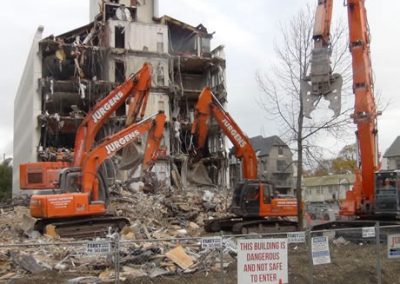 Two Jurgens excavators demolishing the earthquake-damaged Scales House beside a sign reading "This building is dangerous and not safe to enter"