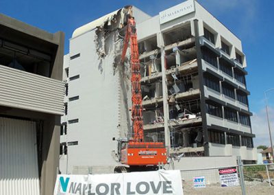 High reach excavator demolishing a multi-storey office block behind a Naylor Love site banner