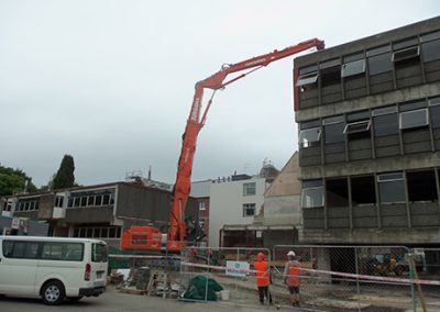 High reach excavator working on a multi-storey concrete building while workers watch from the cordon